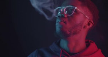 Karanlık arka planda neon ışıkta elektronik sigara içen dalgın bir Afrikalı Amerikalı. Buhar makinesi ve insan konsepti.