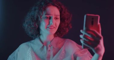 Neşeli genç bir kadın internet üzerinden akıllı telefonuyla neon ışıkta dans ediyor. Gençlik konsepti için sanal buluşma ve gece hayatı.