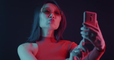 Asyalı kadın, arka planda neon ışığı altında akıllı telefon kamerasıyla selfie çekiyor. Fotoğraf ve modern yaşam tarzı kavramı.