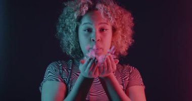 Afrika kökenli Amerikalı tatlı bir kızın konfeti üflerken ve arka planda neon ışıkta gülümserken yavaş çekimde portresi. İyi ruh hali ve kutlama konsepti.