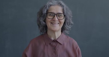 Gri arkaplanda kameraya bakıp gülümseyen mutlu yaşlı bir kadının yavaş çekim portresi.