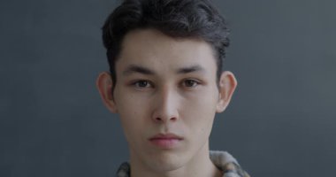 Ciddi bir Asyalı adamın yakın çekim portresi. Gri arka planda ciddi yüz ifadesi olan kameraya bakıyor. İnsanlar ve duygu kavramı.