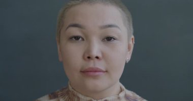 Ciddi bir Asyalı kadının gri arka planda sakin bir yüz ifadesiyle kameraya bakışının yavaş çekim portresi. İnsanlar ve duygu kavramı.