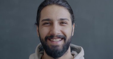 Orta Doğulu neşeli bir adamın gri arka planda kameraya bakıp gülen yavaş hareket portresi. İnsanlar ve olumlu duygu kavramı.