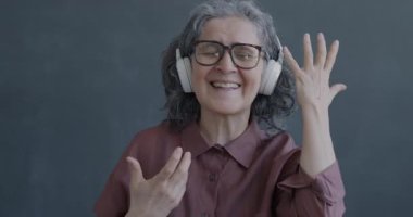 Yavaş çekimde dans eden mutlu son sınıf kadınının gri arka planda kulaklıkla müzik dinlemesi. İnsanlar ve eğlence konsepti.