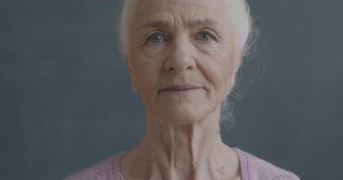Siyah arkaplanda duygusuz bir şekilde kameraya bakan düşünceli yaşlı bir kadının yavaş çekim portresi. İnsanlar ve yüz ifadesi kavramı.