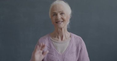 Siyah arka planda kameraya bakarak el sallayan ve gülümseyen neşeli yaşlı bayanın ağır çekim portresi. İnsanlar ve duygu kavramı.