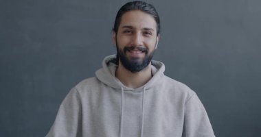 Orta Doğulu neşeli bir adamın gri arka plandaki kameraya el sallayarak gülümsemesinin yavaş çekim portresi. İnsanlar ve ifade kavramı.