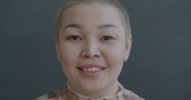 Gri arka planda kameraya gülümseyen neşeli Asyalı genç bayanın yavaş çekim portresi. İnsanlar ve olumlu duygu kavramı.