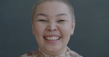 Aşırı neşeli Asyalı kadının gri arka planda eğlenen kameraya bakıp gülen ağır çekim portresi. Mutluluk ve insan kavramı.