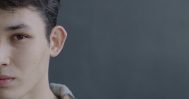 Gri arkaplanda duygusuz bir şekilde kameraya bakan Asyalı genç bir adamın yavaş çekim portresi. İnsanlar ve ifade kavramı.
