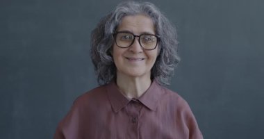 Gri arka planda kameraya bakıp el sallayan neşeli, yaşlı bir kadının yavaş çekim portresi. İnsanlar ve ifade kavramı.