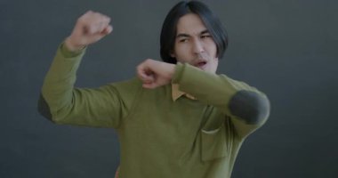 Gri arka planda kameraya bakarak dans eden neşeli Asyalı adamın yavaş çekim portresi. İnsanlar ve eğlence konsepti.