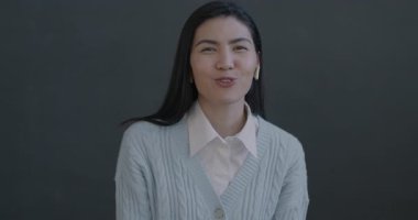 Heyecanlı Asyalı genç bir kadının portresi çevrimiçi video görüşmesi yapıyor. Gri arka planda kulaklık kullanarak el sallıyor. İnsanlar ve iletişim konsepti.