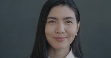 Gri arka planda kameraya gülümseyen güzel Asyalı kadının yavaş çekim portresi. İnsanlar ve olumlu duygu kavramı.