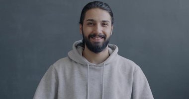 Orta Doğulu mutlu bir adamın gri arka planda gülümseyen el hareketlerini gösteren yavaş çekim portresi. İnsanlar ve olumlu duygu kavramı.