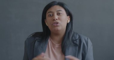 Çevrimiçi video konuşması yapan neşeli genç bir kadının portresi. Kablosuz kulaklıkla siyah arkaplandaki kameraya bakarak işaret ediyor..