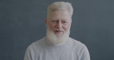 Online video görüşmesi yapan yaşlı bir adamın portresi siyah arkaplandaki kablosuz kulaklıkları kullanarak kameraya bakıp işaret ediyor..