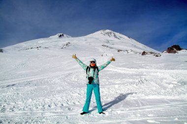 Girl skier at the mountain Elbrus. Smiling happy woman standing in a snowy mountain landscape in Caucasus stretching her arms. In background two summit of Elbrus. Adventure winter extreme sport clipart