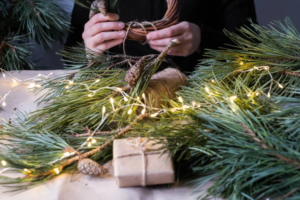 Making rustic Christmas wreath. Female hands on background of  wooden table with pine tree branches, cones, lights.Holiday preparations