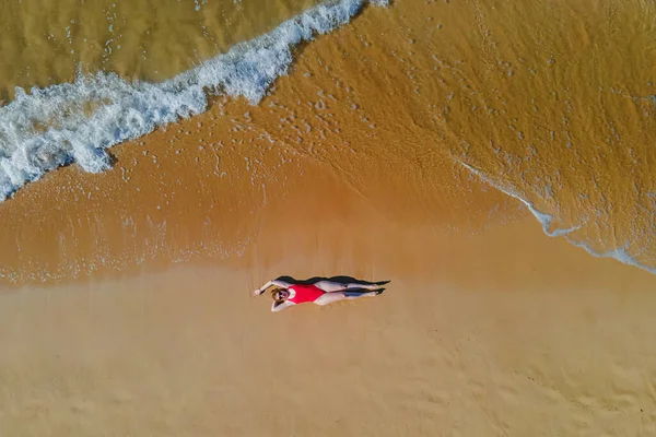 Woman in red swimsuit lying on the beautiful sandy beach with ocean waves, drone aerial view.