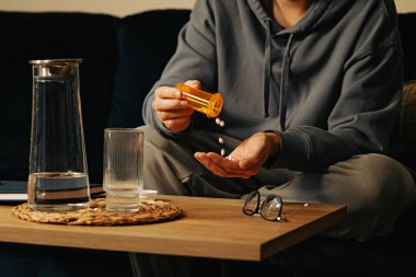 Tanınmayan bir adam elinde turuncu plastik şişeden tabletleri döküyor, geceleri kanepede oturuyor.