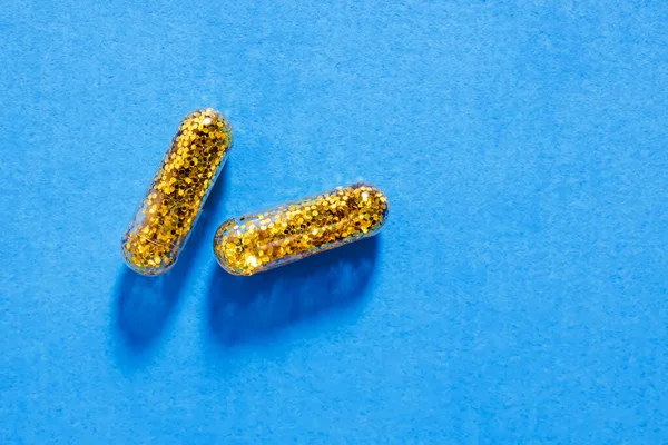 Two pills filled with gold glitter on blue background. Creative concept, drugs, pills