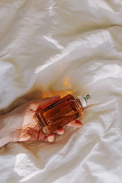 Γυναικείο Χέρι Που Κρατά Ένα Μπουκάλι Άρωμα Στο Λευκό Κρεβάτι — Φωτογραφία Αρχείου