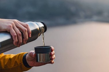 Arka planda manzaralı termostan sıcak çay döken erkek ellerinin yakın çekimi.