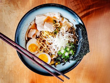 Dilimlenmiş tavuk, yumurta, yosun ve ahşap bir masada çubukları olan yeşil soğanlı ramen kasesi..