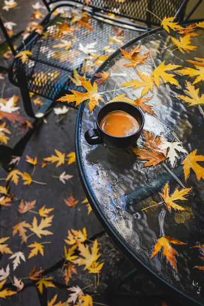 Cup of coffee standing on the patio table after the fall rain with lots of fallen maple yellow leaves
