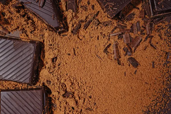 stock image Pieces and shaves of dark bitter chocolate scattered on cocoa powder background, top view