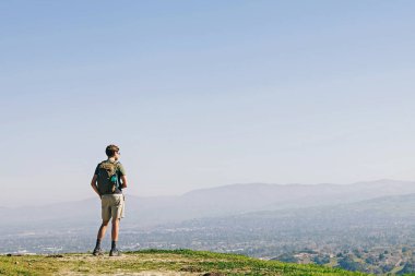 Sırt çantalı bir insan, açık mavi gökyüzünün altında dağları olan geniş bir manzaraya bakan çimenli bir tepenin üzerinde durur..