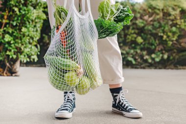 Taze sebze ve meyvelerle dolu, günlük giysiler ve spor ayakkabılar giyen, yeniden kullanılabilir bir ağ çantası taşıyan kişi..