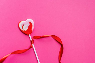 Renkli kalp şeklindeki lolipop parlak pembe bir arka planda duruyor ve çubuğun etrafına kırmızı bir kurdele bağlanmış.