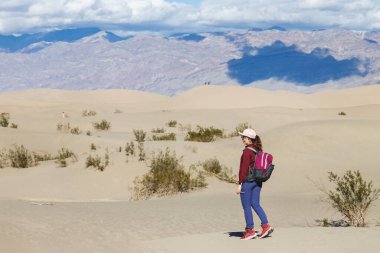 Yürüyüşçü, çöl manzarasında parlak bir gökyüzünün altında geniş kum tepelerini keşfeder.