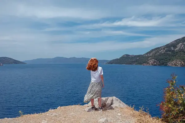 Bir kadın yaz günü sakin mavi denize bakan kayalık bir uçurumda duruyor.