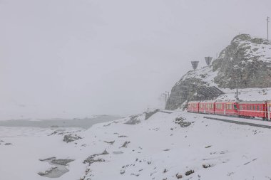 Dağlık bir patika boyunca ilerleyen kırmızı bir trenle karla kaplı bir manzara.