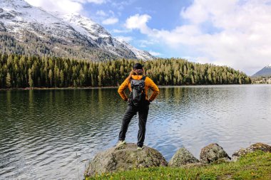 Bir yürüyüşçü sakin bir gölün kenarındaki bir kayanın üzerinde duruyor, dağların ve yemyeşil ormanların manzarasına bakıyor, doğanın huzurunun tadını çıkarıyor..
