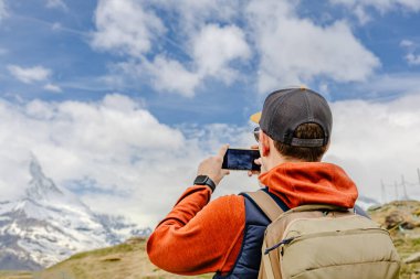 Şapka ve sırt çantası takan bir turist, akıllı telefon kullanarak karla kaplı bir dağın fotoğrafını çekiyor..