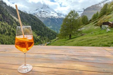 Bir bardak portakal ve pipetle birlikte bir bardak portakal kokteyli bir dağ manzarasına tepeden bakan ahşap bir masada oturur.