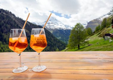 İki bardak turuncu kokteyl, çarpıcı bir dağ manzarasına bakan ahşap bir masada oturur. Canlı içkiler ve doğal arka plan rahatlamak için mükemmel bir ortam oluşturuyor..