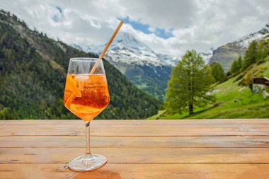 Bir bardak turuncu kokteyl, karlı dağların manzaralı arka planı ve bulutlu bir gökyüzünün altında yemyeşil bir zeminde, ahşap bir masada oturur. Ayarlar, sakin bir açık hava rahatlama noktası olduğunu gösteriyor..