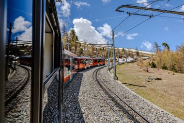 Canlı turuncu bir tren, berrak mavi bir gökyüzünün altındaki resimsel dağlık bir arazide kavisli bir yolda ilerliyor..