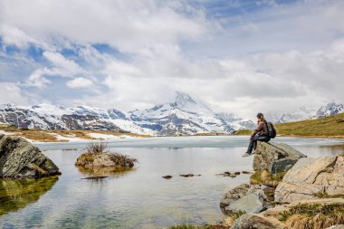 Bir insan kayanın üzerinde oturmuş, karla kaplı bir dağ ve sakin bir gölün huzurlu manzarasının tadını çıkarıyor. Sahne doğanın güzelliğini ve ıssızlığın huzurunu yakalıyor..