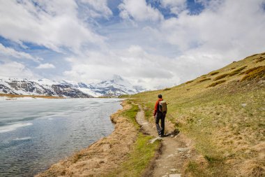 Karla kaplı dağlarla ve bulutlu gökyüzünün altındaki yemyeşil tepelerle çevrili, manzaralı bir yolda yürüyen bir yürüyüşçü.