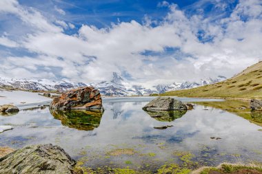 Sakin bir alp gölü etrafındaki karlı dağları ve parlak mavi gökyüzünün altındaki kayaları yansıtır..