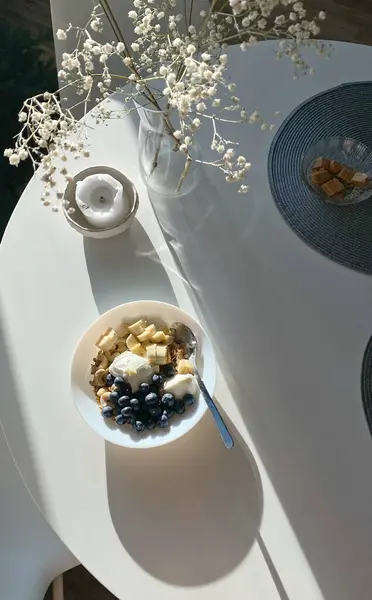上图是装饰有家居装饰的白色餐桌 还有一碗带早餐水果的麦片 光影兼有 — 图库照片