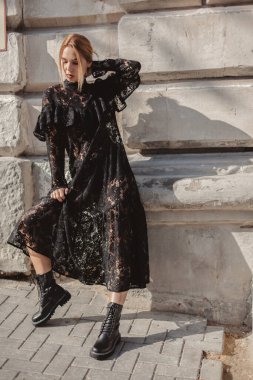 Açık havada, siyah dantelli maksi elbise giyen zarif bir kadının yaz portresi ve şehir sokaklarında poz veren sonbahar deri çizmeleri. Doğal güneş ışığı. Modelin arkasındaki şehir binaları.