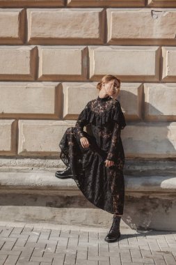 Açık havada, siyah dantelli maksi elbise giyen zarif bir kadının yaz portresi ve şehir sokaklarında poz veren sonbahar deri çizmeleri. Doğal güneş ışığı. Modelin arkasındaki şehir binaları.